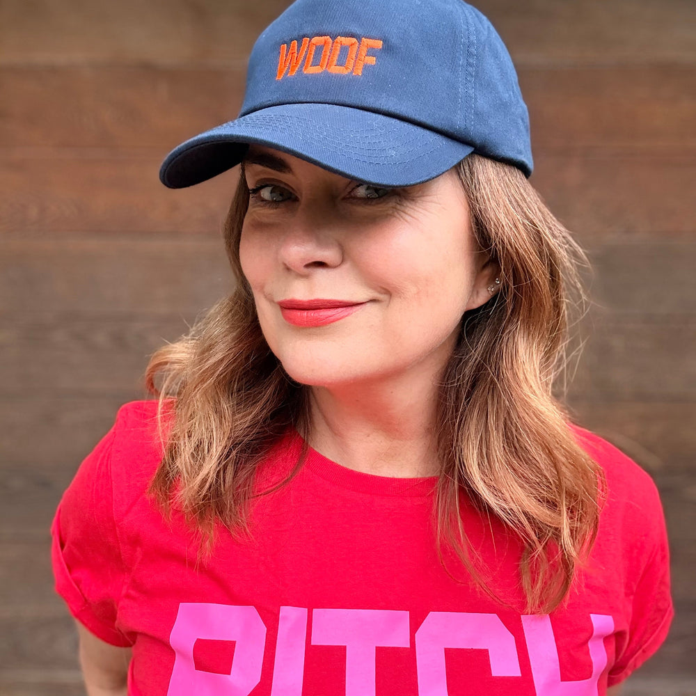Person wearing a blue cap with 'WOOF' and a red shirt with 'BITCH' text against a wooden background